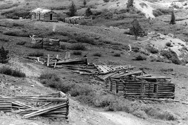 One b/w photograph looking down Aspen Avenue (Main street) of the Ghost Town of Independence. In the foreground of the photograph on the right side of the street is a cabin with walls still standing which can be seen from the side. Along the same side is a row of collapsed structures. On the left side of the road is a single structure which has at least three walls still standing but no roof. In the background of the photograph there are two standing cabins standing parallel to each other. The cabin on the left has a partial roof still intact while the right cabin appears to have wood still attached to the roof. Published in the Aspen Times July 3, 1980 on page 1-b.