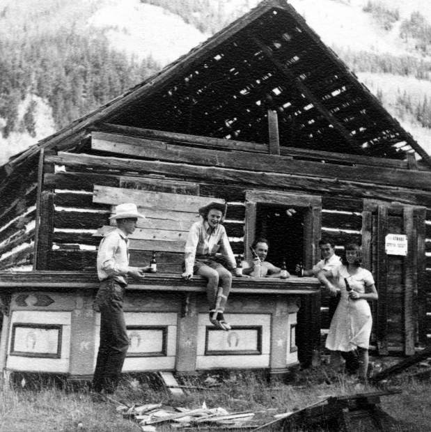 One b/w photograph of the the Blue Mirror Saloon in Ashcroft. A wooden bar sits outside and five people are surrounding it (left to right-unidentified man, Joan Trumbull, unidentified man, unidentified man, and Maud Banks). Joan is sitting on the bar and has a splint on her left leg, and all of them are drinking a beer. The saloon is falling apart, and there is wood scattered around on the ground. 1948