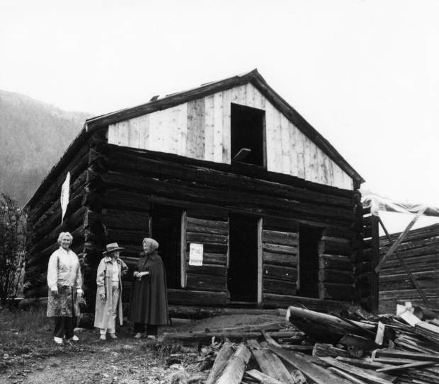 One b/w photograph of three unidentified women standing in front a building at the ghost town of Independence. The building is being restored, and the upper section of the building is constructed of new, lighter wood, and there are several pieces of dark wood lying in front of the building. The women were at Independence to celebrate the 